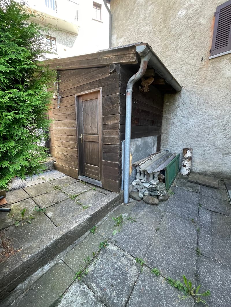 Wooden shed with a stone wall, a wooden door, and a chimney pipe. There is a stack of firewood next to the shed.