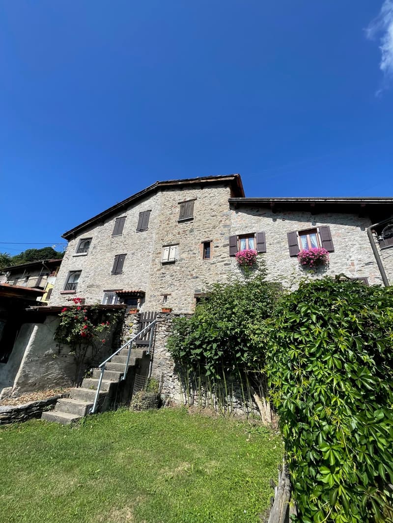 Traditional stone building, wooden shutters, pink flowers in window boxes, stone wall and steps in front, well-maintained garden