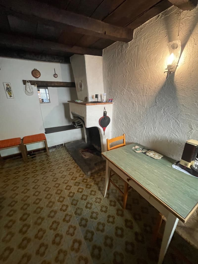 The image shows a rustic kitchen with a fireplace, wooden furniture, and patterned floor tiles. There are various cooking utensils and appliances visible, indicating this is a functional kitchen space.