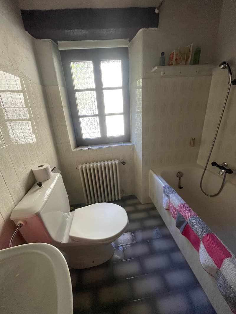 Small bathroom with a toilet, sink, and shower. The room has a window with decorative glass panes and a radiator. There is a shelf above the sink and a towel hanging on the wall.