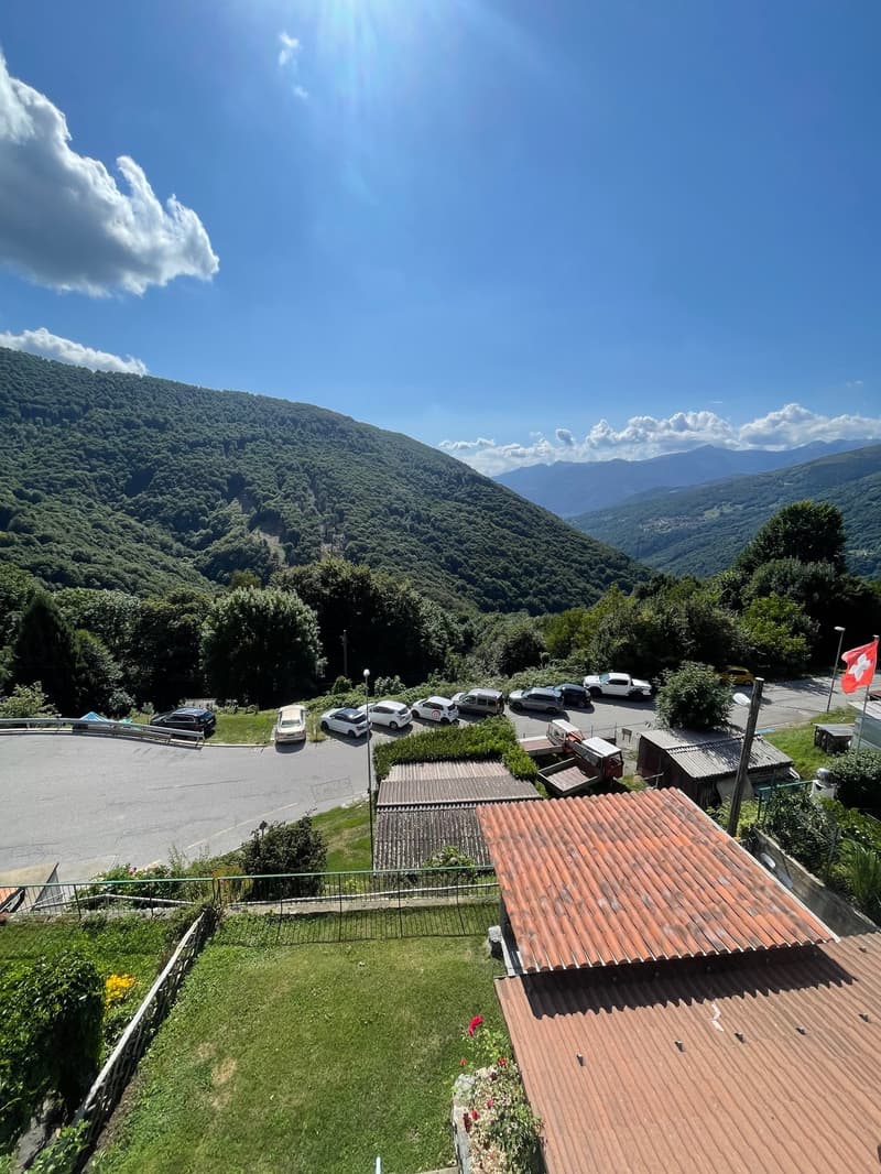 The image shows a scenic mountain landscape with lush green forests covering the hills. There is a parking lot visible in the foreground, surrounded by vegetation. The sky is clear and blue, with some clouds visible. The overall scene appears to be a pict