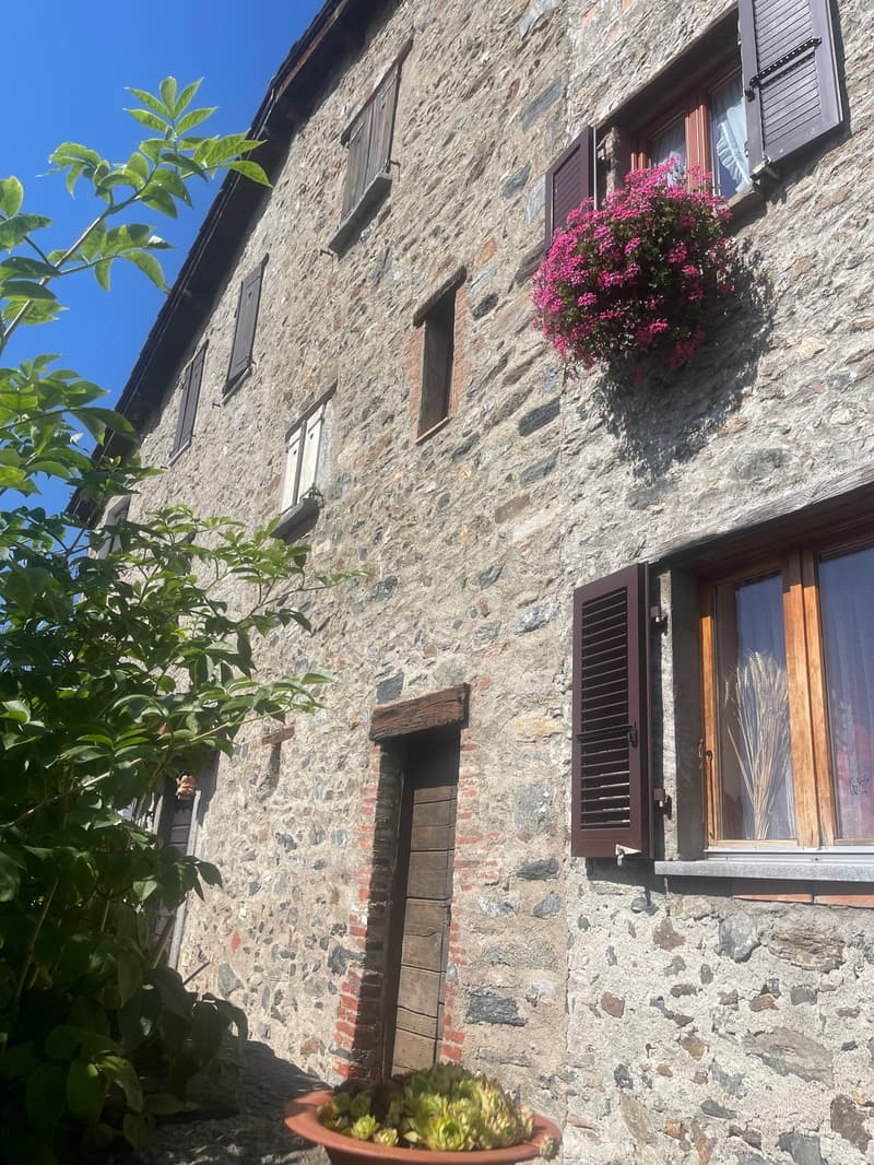 Old stone building with wooden shutters, flower boxes on the balcony, and lush greenery surrounding the entrance.