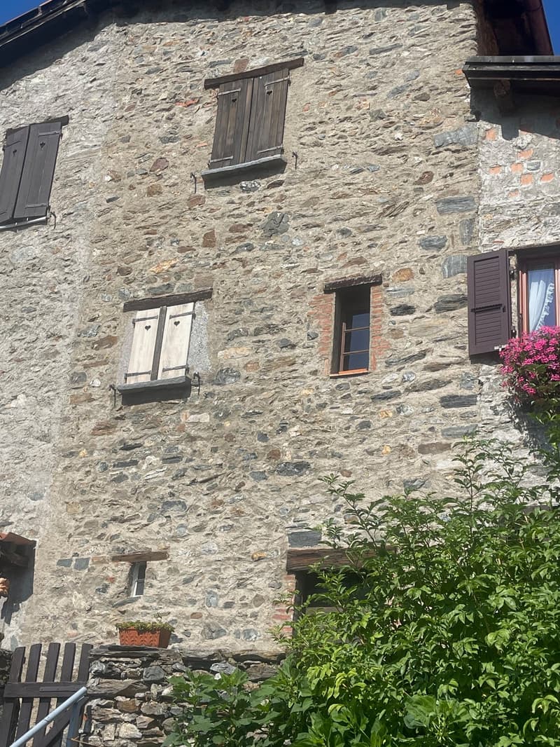 Old stone building with wooden shutters and windows, surrounded by lush greenery.