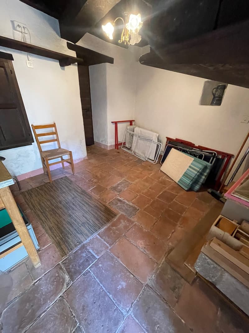 The image shows a room with a tiled floor, wooden beams on the ceiling, and a chandelier. There is a wooden chair in the corner, and various items and furniture stored in the room, including what appears to be a fireplace.