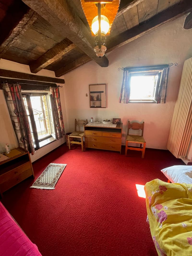Red carpet, wooden furniture, bed with floral sheets, dresser with sink, wooden chairs, radiator, picture frame, lamp, two windows with curtains.
