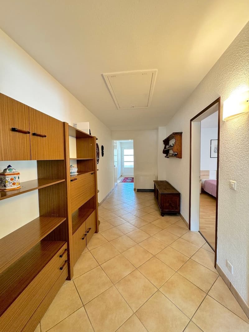 tiled floor, wooden cabinets, white walls, wooden chest, wooden shelf, light fixture, open doorway to bedroom, rug, bed