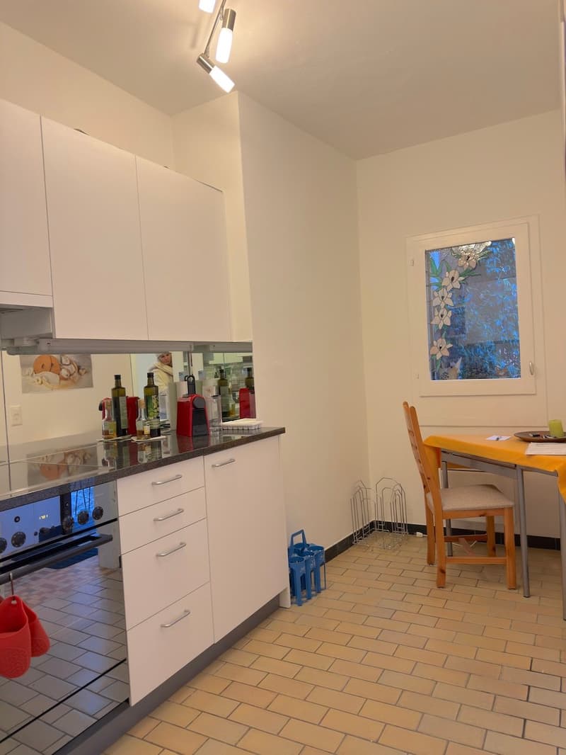 white cabinets, stainless steel appliances, granite countertop, tiled flooring, window with glass design, dining table and chairs, wine bottles and other items on counter, red slippers
