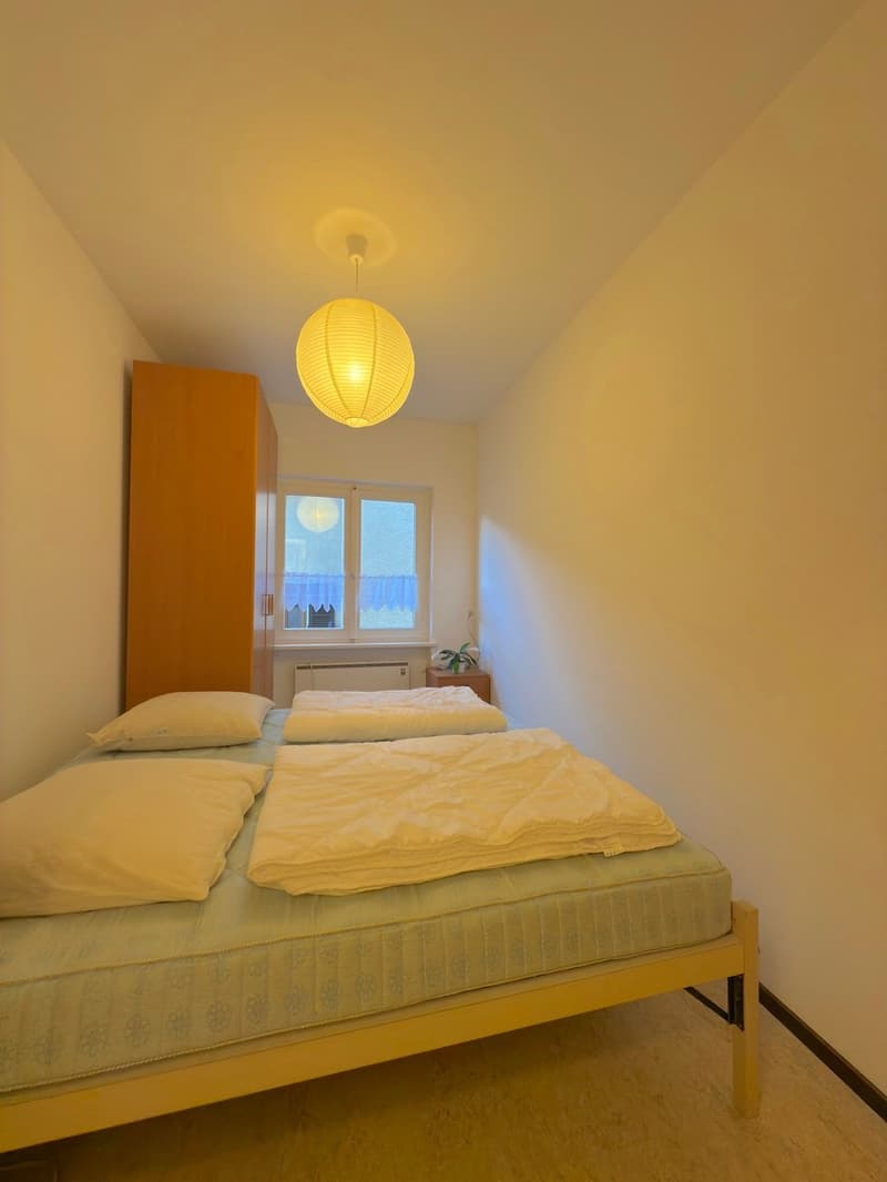 Three beds, wooden floor, white walls, wardrobe, window, lamp, nightstands, pillow