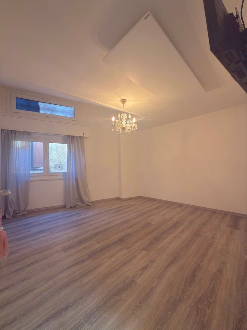 Empty room with wooden floors, chandelier, windows with curtains, and a mounted TV.