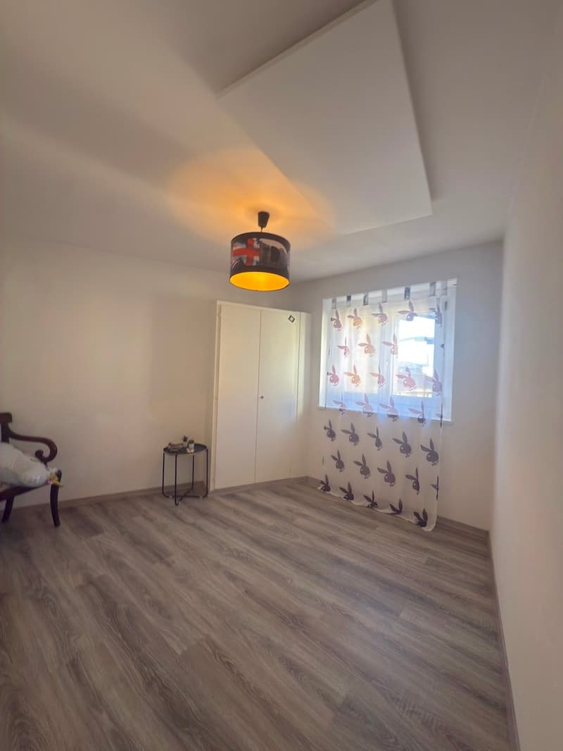 An empty room with a white wall and wooden flooring. The room features a white door and a white window with a curtain. There is a small round table with a potted plant and a lamp in the middle of the room.