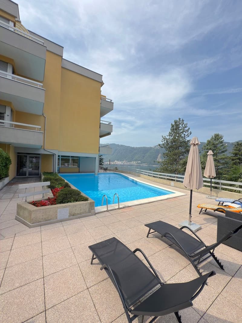 An apartment building with a swimming pool, lounge chairs, and umbrellas on the terrace, overlooking a mountain and a lake.