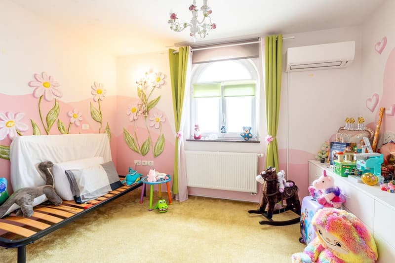 Children's room with pink and white walls, floral wall art, bed, toys, rocking horse, radiator, window with curtains, air conditioner, chandelier