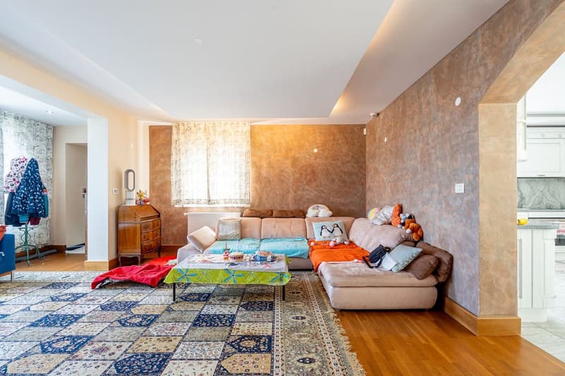 living room with a couch, coffee table, rug, wooden floor, wall with a window and curtains, wooden cabinet, and a kitchen area