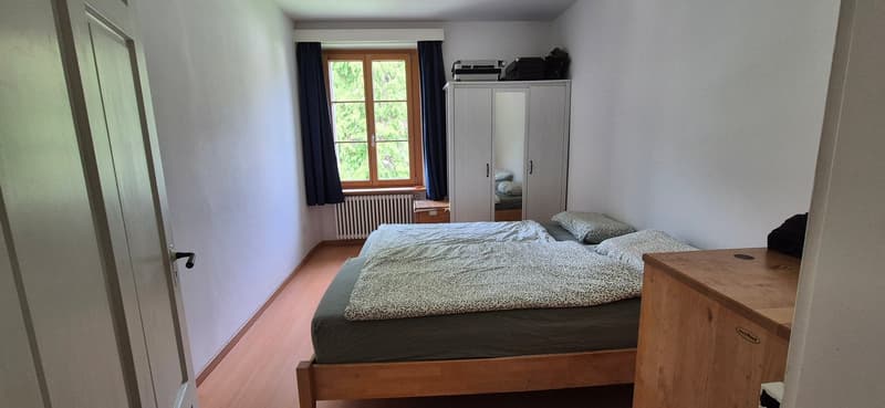Bedroom with a double bed, wooden floor, white walls, window with curtains, wardrobe, and a wooden desk.