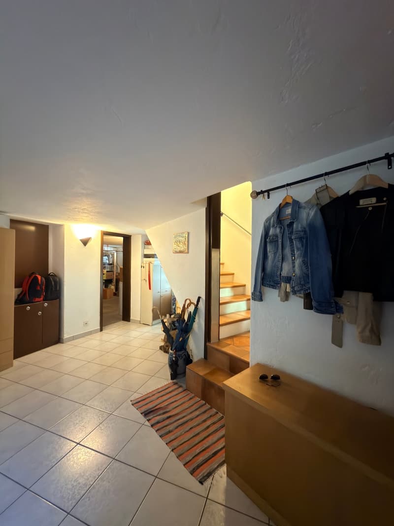 Entrance hall with white tiles, wooden bench, hanging clothes, umbrellas, picture frame, cabinets, and a wall-mounted light.