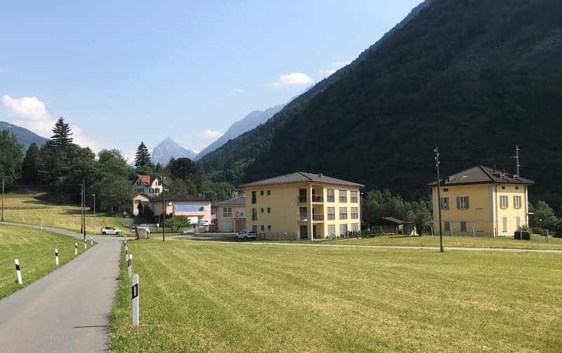A large two story apartment complex in a green valley, several other homes behind it, surrounded by grass fields and mountains