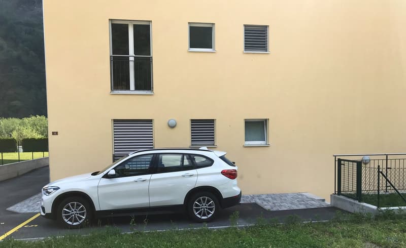 The property has a white SUV parked in front of a two-story building with a garage area. The building features a yellow exterior with a few windows. The parking area is paved with gray bricks, with a fence and a small lawn.