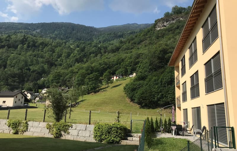 A yellow building with windows and awnings, outdoor seating, fenced yard, a stone wall, and a scenic view of the mountains and green fields.