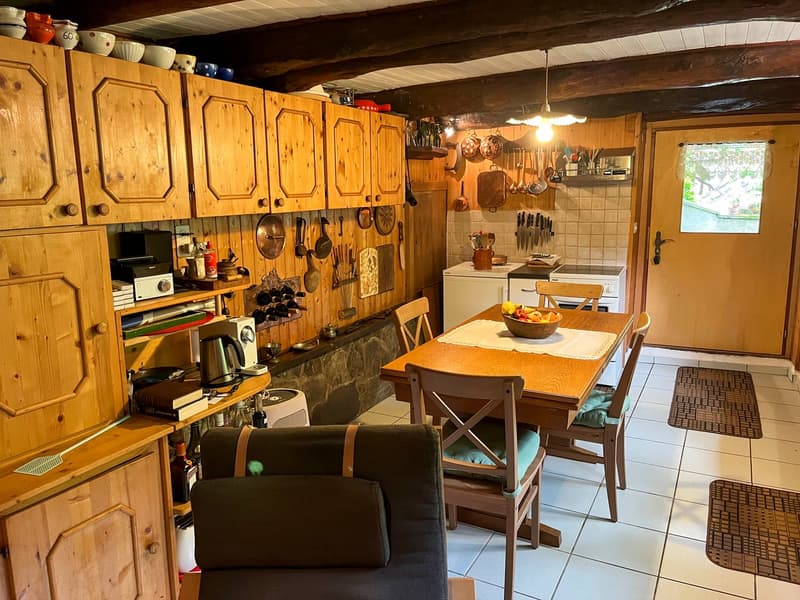 Dining area with a wooden dining table and chairs, wooden cabinets and shelves, tiled floor, kitchen counter with stove and appliances, and hanging pots and pans.