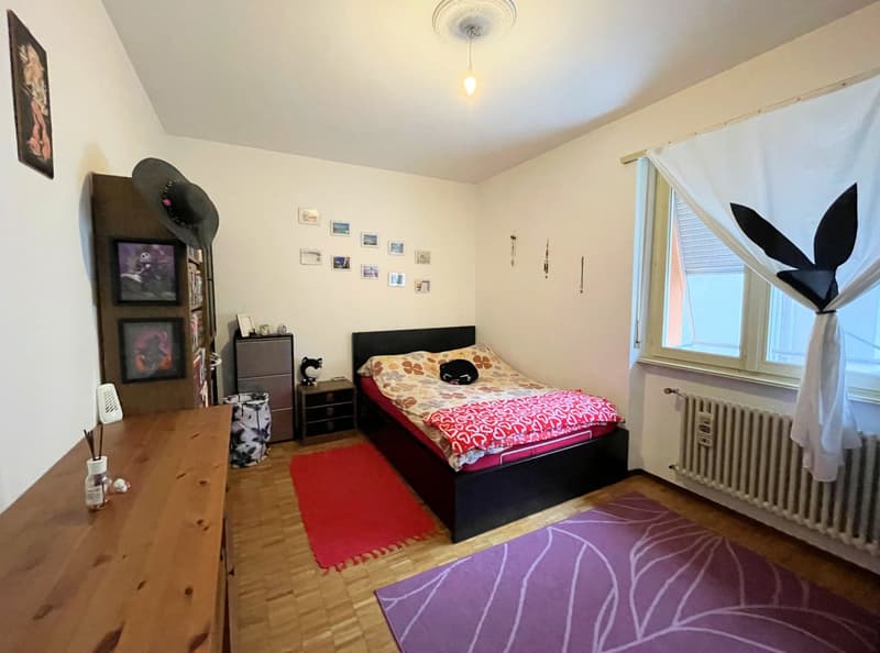 Bedroom with hardwood floor, white walls, bed with colorful bedding, dresser, shelving unit, and framed artwork on the walls.