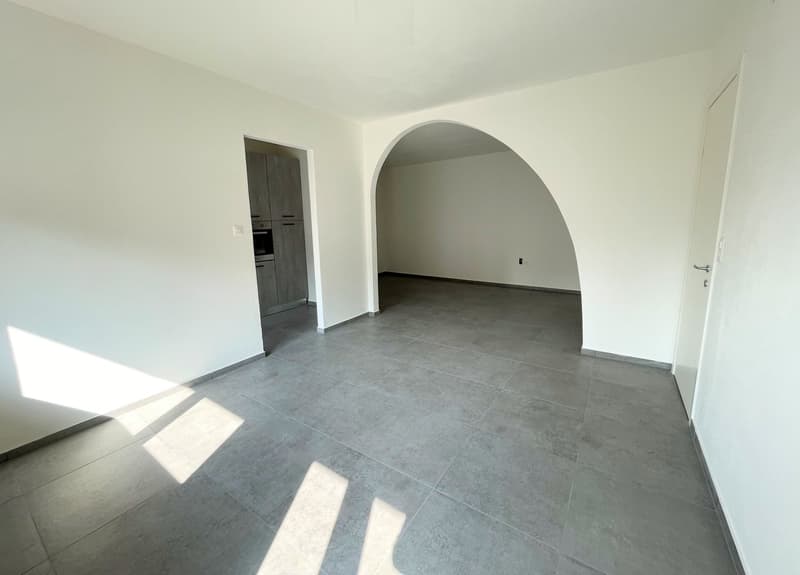 An empty room with gray tiled flooring, white walls, and a small archway on the left side leading to another room. An oven and cabinets are visible in the room through the archway. A power outlet is seen on the wall on the right.