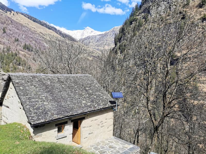 Stone house with solar panel on the roof, wooden door, surrounded by trees and mountains