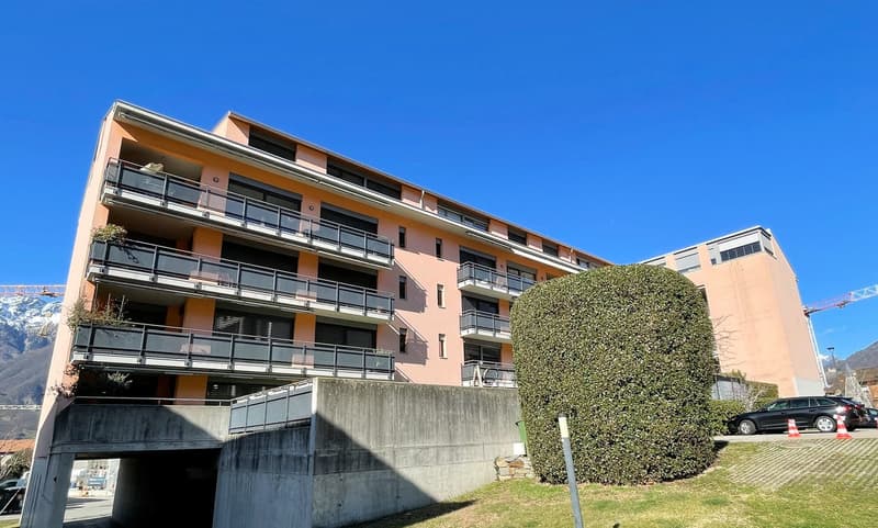 A multi-story residential building with multiple balconies, surrounded by greenery and parking spaces, set against a backdrop of mountains.