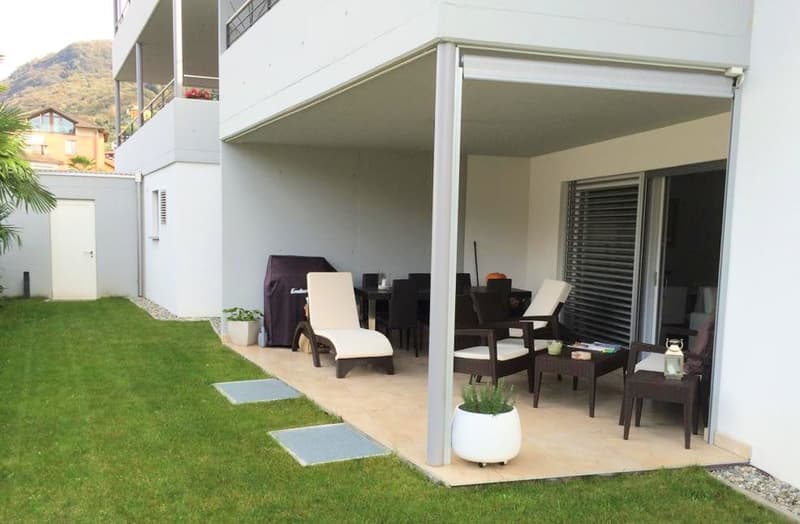 covered outdoor patio, seating area with white lounge chairs, round tables, garden with green grass, potted plants, and shrubs
