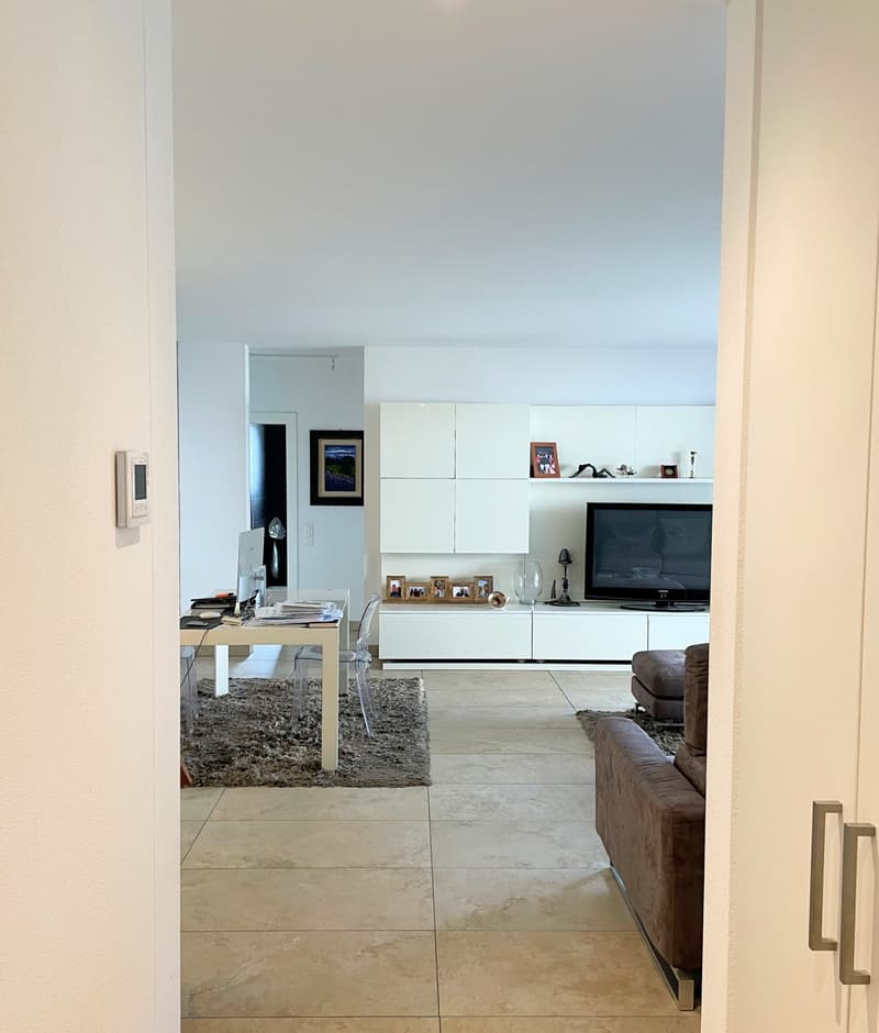 large room with white walls, beige tiles, sofa, coffee table, TV, desk, chairs, rug