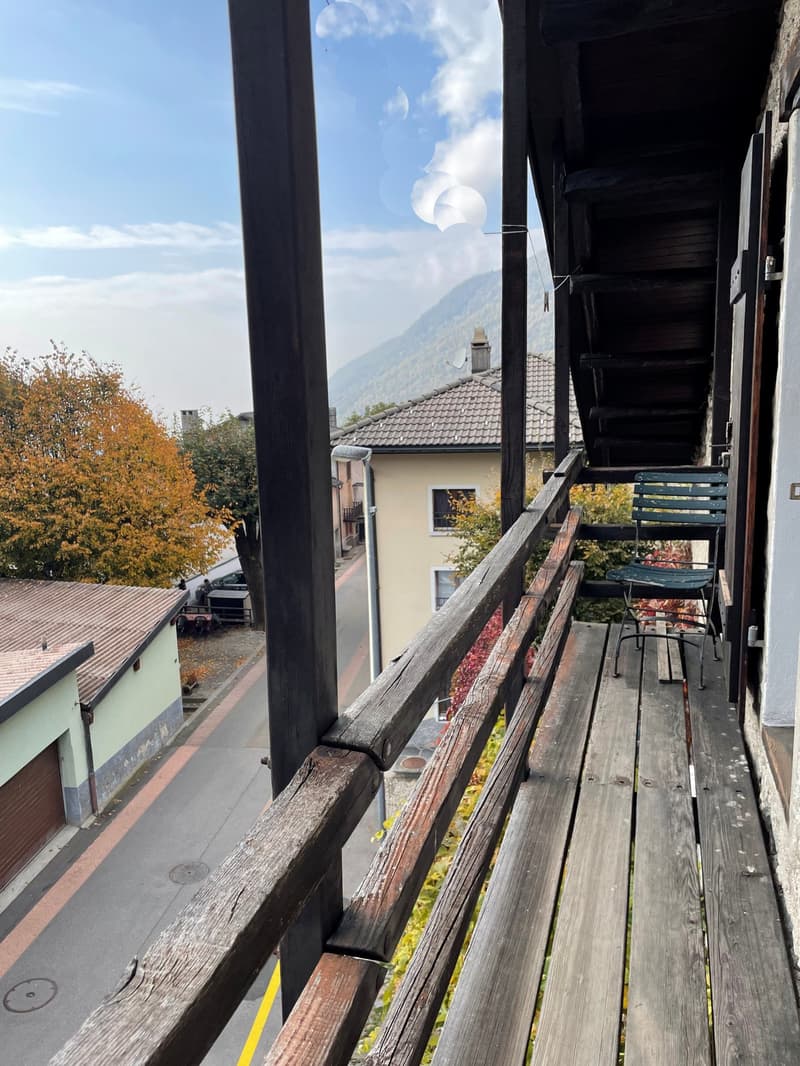 A balcony with wooden railings, a wooden floor, a folding chair, and a distant view of a house and mountains