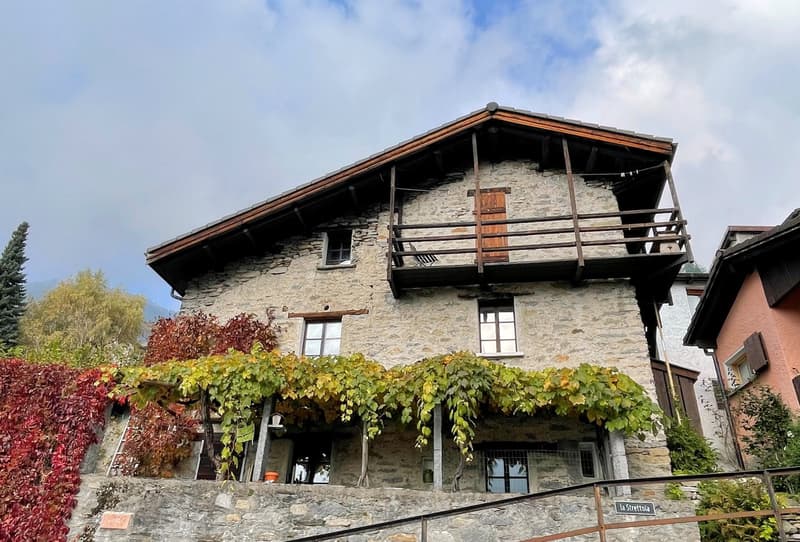 Stone house, brown roof, wooden balcony, raised ground floor, red leafy plants