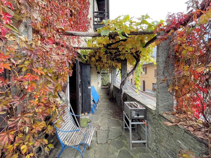 stone pathway, arched stone wall, wooden chair with blue frames, wooden archway with plants, grill on metal stand