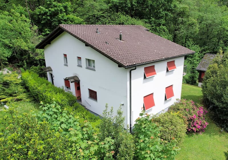 2-story house, white exterior, brown roof, situated on a hillside with greenery and trees, small balcony on the upper floor