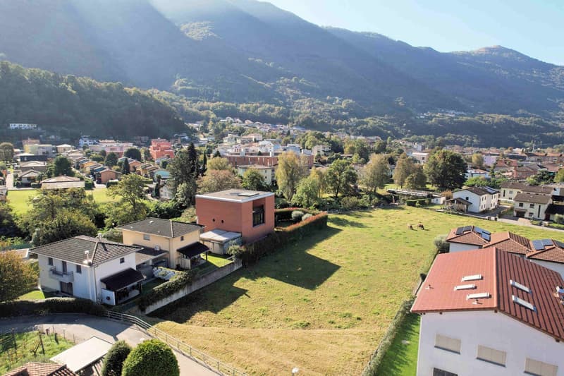 Aerial view of a town with several houses, some with solar panels, a large grassy area, and mountains in the background.