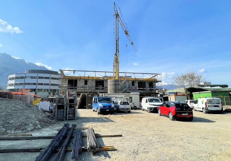 Construction site with a crane, several vehicles parked, building under construction, mountains in the background