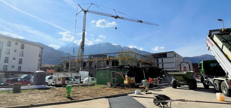 Construction site with a crane, machinery vehicles, and workers in progress. Buildings under construction, mountains in the background.