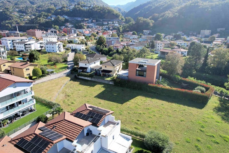 House with solar panels on the roof, surrounded by a large green field, multiple houses in the background, mountains in the distance
