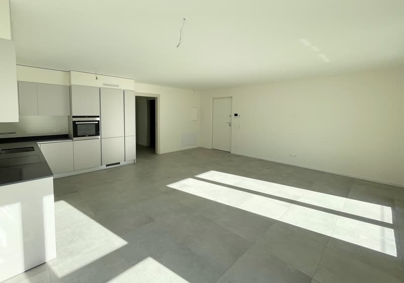 Empty room with white walls, gray floor, kitchen area with cabinets, oven, and countertop, door leading to another room