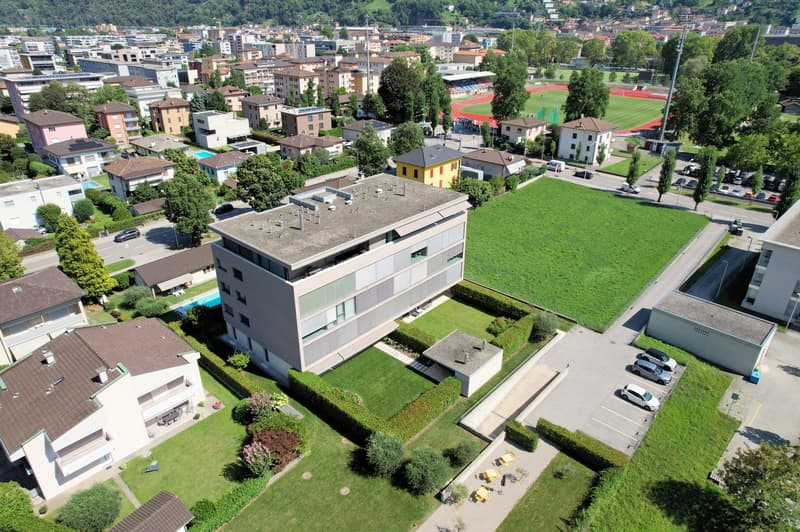 Aerial view of a residential building with a parking lot, surrounded by other buildings, trees, and a green field.