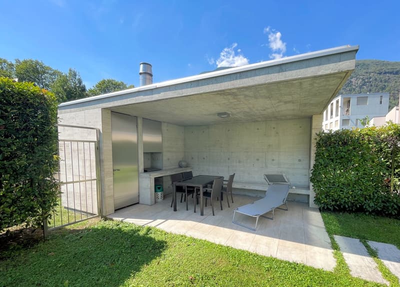 Outdoor dining area with table and chairs, metal gate, concrete walls, small garden, green grass, plants, and trees