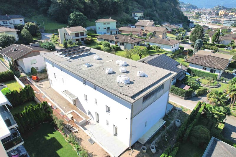 Aerial view of a modern white building with solar panels on the roof, surrounded by other houses and greenery.