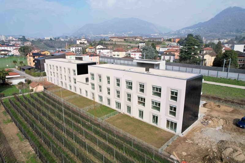 A large white building with many windows, surrounded by a fence, with a vineyard in front and a mountain in the background.
