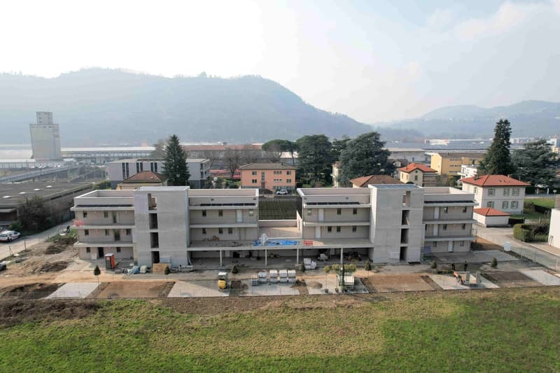 Newly constructed multi-story building, solar panels on the roof, swimming pool in front, surrounded by trees and grass