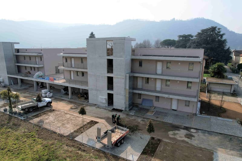 A multi-story building with balconies, under construction, surrounded by trees and mountains