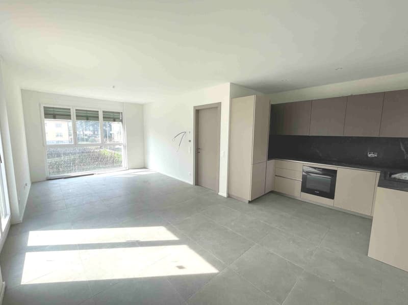 Empty room with white walls, gray tiled floor, large windows, kitchen area with oven and cabinets