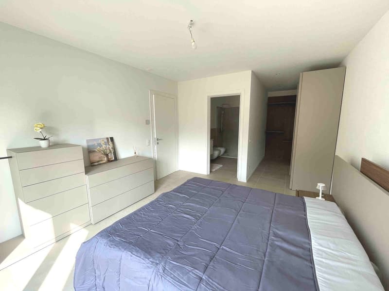 Bedroom with a double bed, white walls, white dresser, white nightstand, white lamp, white flower vase, picture frame, and a bathroom with a toilet and shower.