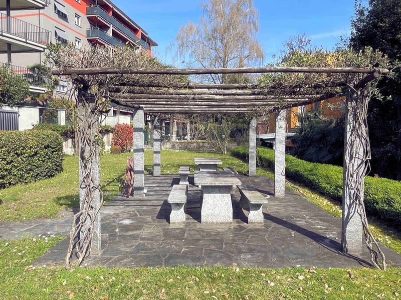 Outdoor seating area with stone benches and tables under a pergola covered with vines, surrounded by a grassy lawn and hedges, with a building in the background.