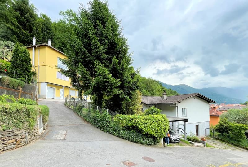 Two houses, one yellow and one white, with a car parked in front of the white house, surrounded by greenery and mountains in the background.