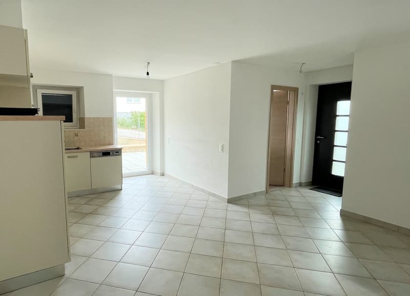 Empty room with white tiles, white walls, kitchen area with sink and dishwasher, glass door leading to outside, wooden door, black door