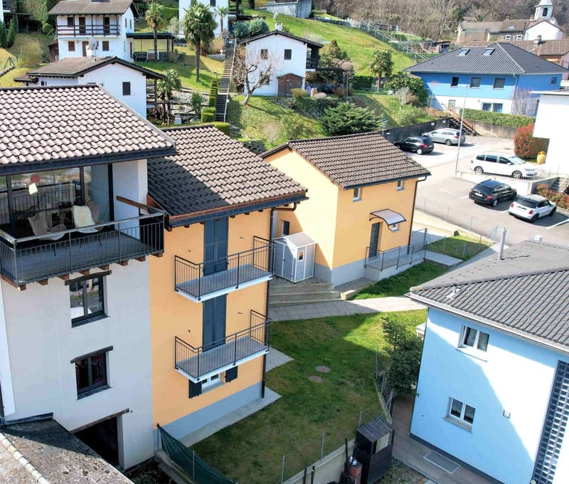 Aerial view of a group of houses, one with a balcony, surrounded by a green hill, with cars parked in the parking lot.